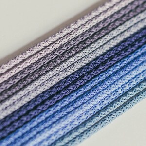 May include: Close-up of a bundle of blue, purple, and gray cords. The cords are tightly woven and arranged in parallel lines, creating a gradient effect from light to dark. The background is a plain, off-white surface.