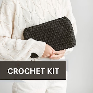 Könnte beinhalten: Eine schwarze gehäkelte Clutch mit Reißverschluss. Die Tasche wird von einer Person gehalten, die einen weißen Pullover und eine weiße Hose trägt. Der Text "CROCHET KIT" ist im Bild sichtbar.