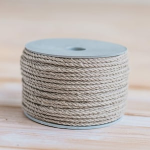 May include: A spool of natural twine wound around a light gray plastic cylinder. The twine is a light beige color and has a rough texture.