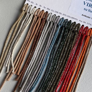 Può includere: Una cartella colori di 18 colori diversi di cordoncino con filo metallico. Il cordoncino è disposto in file, con i colori che vanno dal beige al rosso. La cartella è etichettata con i numeri 10, 44a, 45s, 3s, 4a, 5a, 66a, 11s, 228s, 70s, 207s, 2255, 2095415, 41a, 26s, 217a, 68a e 54a. La cartella include anche il testo "Megztos tušc VIRVU su liurex si" e "Artikulai: 797-3, 79."