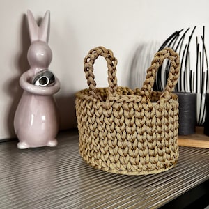 May include: A small, crocheted basket with two handles shaped like bunny ears. The basket is made of a light brown cord and is sitting on a textured surface.