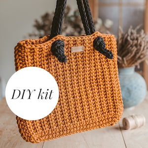 Può includere: Un kit fai-da-te per una borsa a tracolla all'uncinetto. La borsa è realizzata con una corda spessa di colore arancione e ha manici in corda nera. La borsa ha un'etichetta che dice "MONOMY".