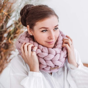 May include: A woman wearing a pink chunky knit infinity scarf. The scarf is made of thick yarn and has a braided texture.