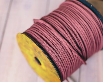 May include: A spool of dusty rose colored cord wound around a yellow plastic spool. A small flower made from the same cord is lying on a white wooden surface.