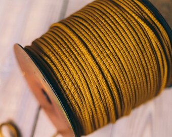 May include: A spool of mustard-yellow rope sits on a light-colored wooden surface. The rope is tightly wound around the spool, with a length of the same rope looped out. The rope has a textured appearance.