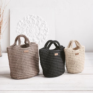 May include: Three crocheted tote bags in different sizes and colors: beige, black, and cream. The bags have leather handles and a small leather tag with the word "handmade" on them.