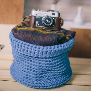 May include: A vintage camera in a brown leather case rests atop a folded patterned sweater and a light blue woven basket. The scene is set on a wooden surface, suggesting a theme of photography and home decor.