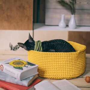 May include: A black cat resting in a yellow crocheted pet bed. The bed is on a wooden surface with books and a sunflower illustration on the cover of one book.