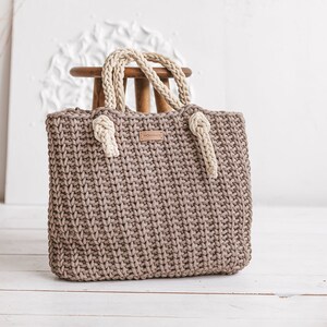 Op de afbeelding: Een bruine gehaakte tote bag met cr&egrave;mekleurige touwhandvatten. De tas heeft een lederen label met het woord "MONOMOMMY" erop.