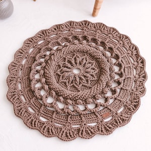 Puede incluir: Una alfombra redonda de crochet marrón con un diseño de mandala. La alfombra tiene varias capas de círculos de crochet con patrones intrincados.