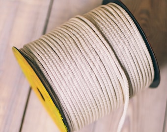 Può includere: Una bobina di corda bianco sporco con una bobina gialla e nera. Un pezzo di corda si estende dalla bobina a un oggetto circolare all'uncinetto. Gli oggetti sono su una superficie di legno chiaro. La corda e l'oggetto all'uncinetto sono beige.