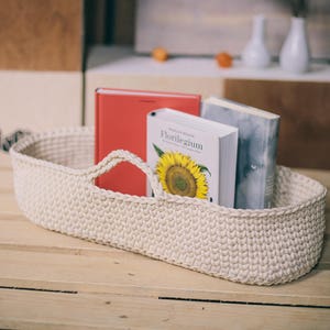 May include: A white crocheted basket with a handle sits on a wooden surface.  Three books are visible in the basket, including one with the title "Florilegium".