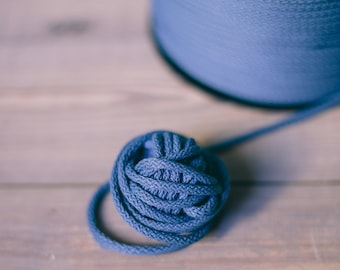 May include: A close-up shot of a ball of blue yarn with a larger spool of yarn in the background. The yarn is a muted blue color, and the ball is tightly wound. The background is a light-colored wooden surface.