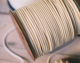Può includere: Primo piano di un rotolo di corda di cotone bianco naturale. La corda è avvolta strettamente attorno a un rotolo di legno con una fascia metallica. Ci sono due rotoli di corda più piccoli sullo sfondo.