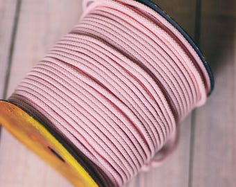 May include: A spool of pink rope sits next to a flower-shaped item made from the same material. The spool has a yellow and brown core. The items are on a light-colored wooden surface.