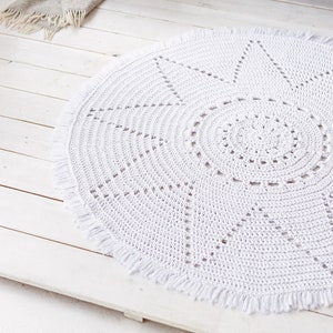 May include: White crocheted round rug with a starburst design and fringe trim. The rug is on a white wooden floor.