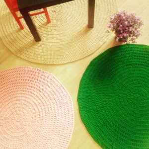 May include: Three round crocheted rugs in different colors: beige, pink, and green. The rugs are arranged on a wooden floor.