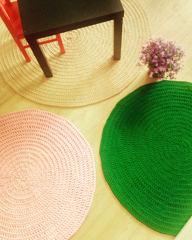 May include: Three round crocheted rugs in different colors: beige, pink, and green. The rugs are arranged on a wooden floor.