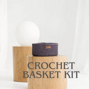 May include: A dark gray crochet basket with a small leather tag, displayed on a wooden pedestal. The text "CROCHET BASKET KIT" is printed on the pedestal. A white sphere and a second wooden pedestal are in the background.