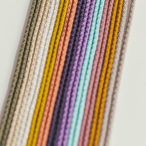 May include: A close-up of a collection of colorful cords in various shades, including beige, yellow, peach, purple, and teal. The cords are arranged in parallel lines against a white background, creating a visually appealing display.