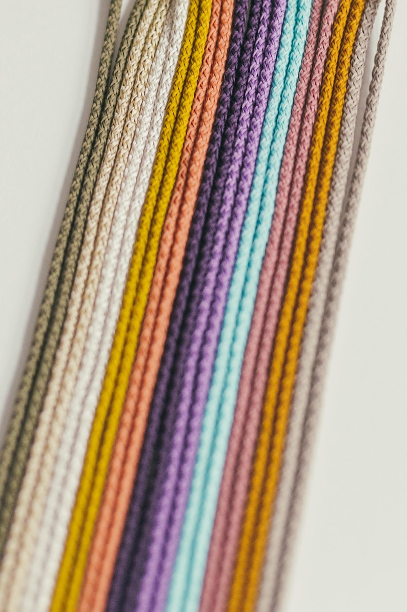 May include: A close-up of a collection of colorful cords in various shades, including beige, yellow, peach, purple, and teal. The cords are arranged in parallel lines against a white background, creating a visually appealing display.
