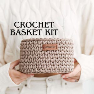Pode incluir: Um kit de cesta de crochê bege neutro. A cesta é cilíndrica com um padrão de ponto texturizado e uma pequena etiqueta de couro que diz "MONOMEY". O texto "CROCHET BASKET KIT" é exibido acima da cesta.