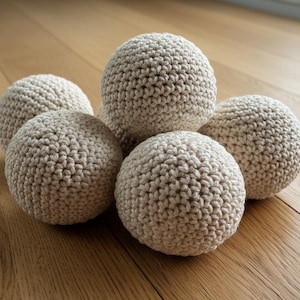 May include: Five beige crocheted spheres of varying sizes are arranged on a wooden surface. The spheres have a textured, handmade appearance, with visible stitch patterns. The neutral color palette and tactile quality suggest a focus on natural materials.