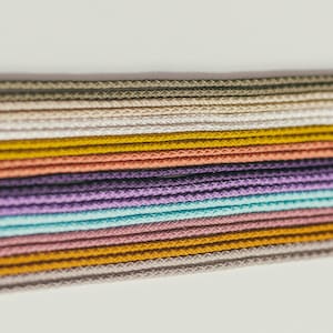 May include: Close-up of a collection of colorful cords in various shades, including beige, yellow, coral, purple, and teal. The cords are tightly woven and arranged horizontally, creating a textured pattern. The image has a soft, natural lighting.