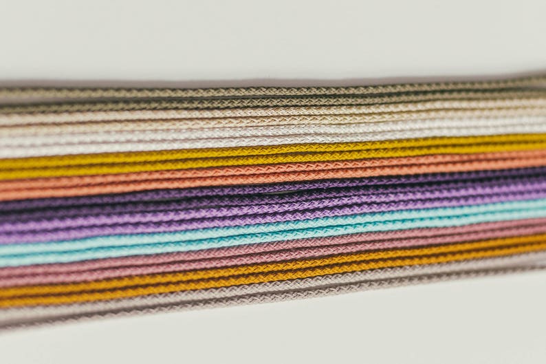 May include: Close-up of a collection of colorful cords in various shades, including beige, yellow, coral, purple, and teal. The cords are tightly woven and arranged horizontally, creating a textured pattern. The image has a soft, natural lighting.