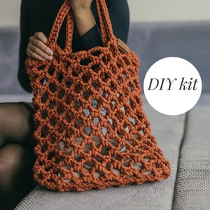 Pode incluir: Um kit de bricolage para uma bolsa de compras de crochê laranja. A bolsa é feita com um fio grosso semelhante a uma corda e tem uma trama larga e aberta. A bolsa está sobre um cobertor cinzento.