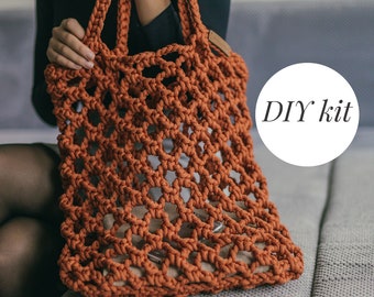 May include: A DIY kit for a crocheted orange tote bag. The bag is made with a thick rope-like yarn and has a large, open weave. The bag is sitting on a gray blanket.