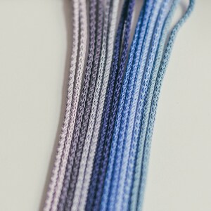 May include: Close-up of a collection of ombre-dyed cords in shades of purple and blue. The cords are tightly woven and arranged in parallel lines, creating a gradient effect. The cords are likely used for crafting or jewelry making.