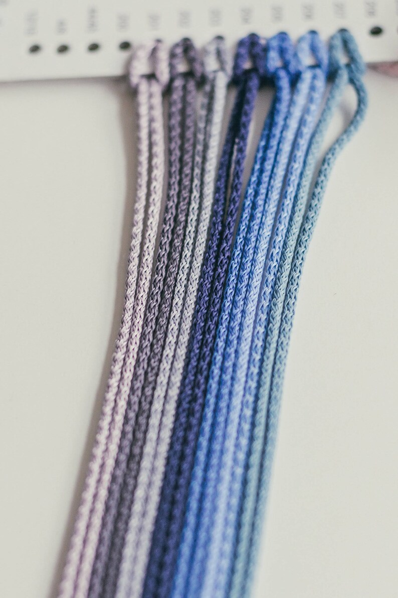 May include: Close-up of a collection of ombre-dyed cords in shades of purple and blue. The cords are tightly woven and arranged in parallel lines, creating a gradient effect. The cords are likely used for crafting or jewelry making.