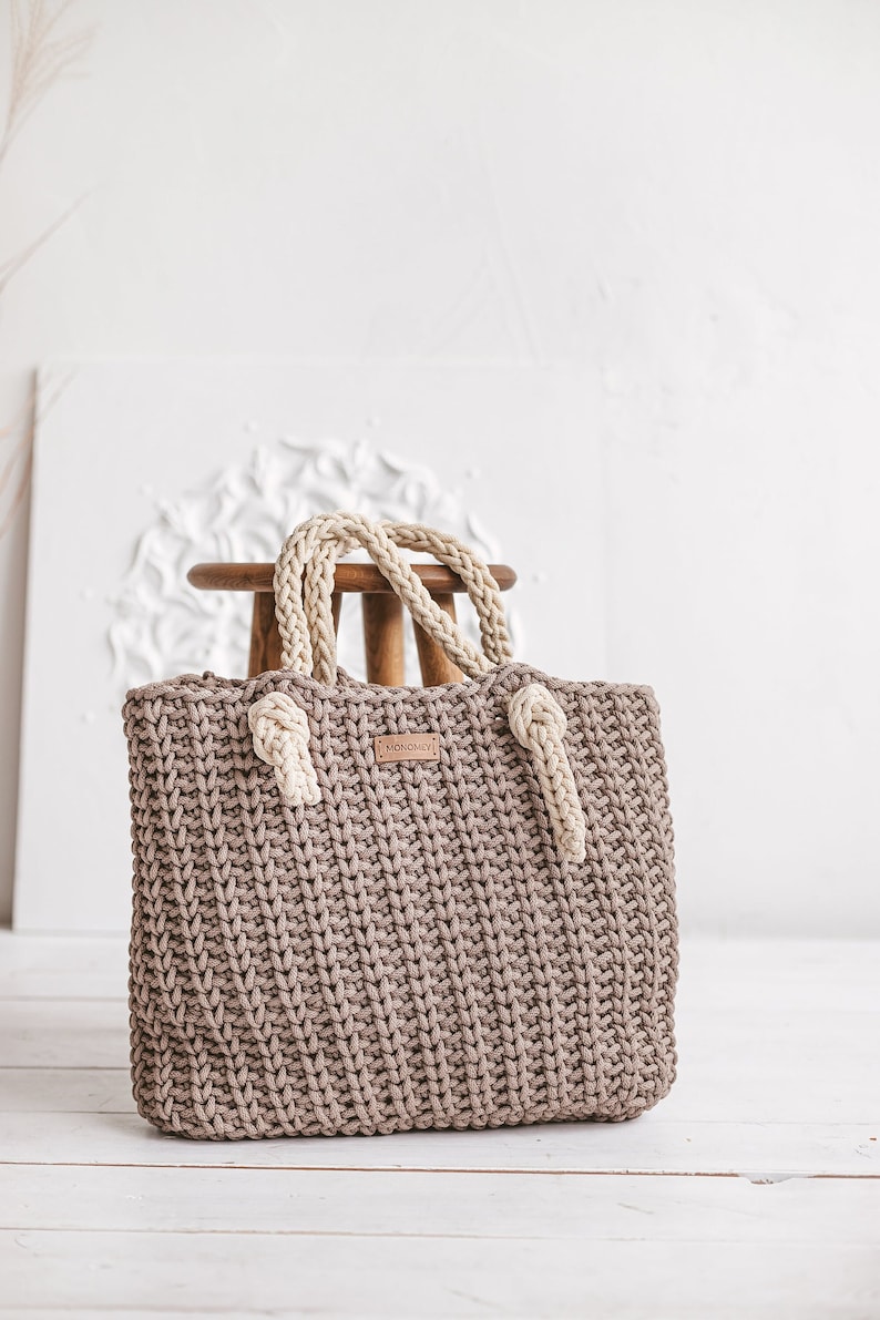Op de afbeelding: Een bruine gehaakte tote bag met cr&egrave;mekleurige touwhandvatten. De tas heeft een lederen label met het woord "MONOMOMMY" erop.