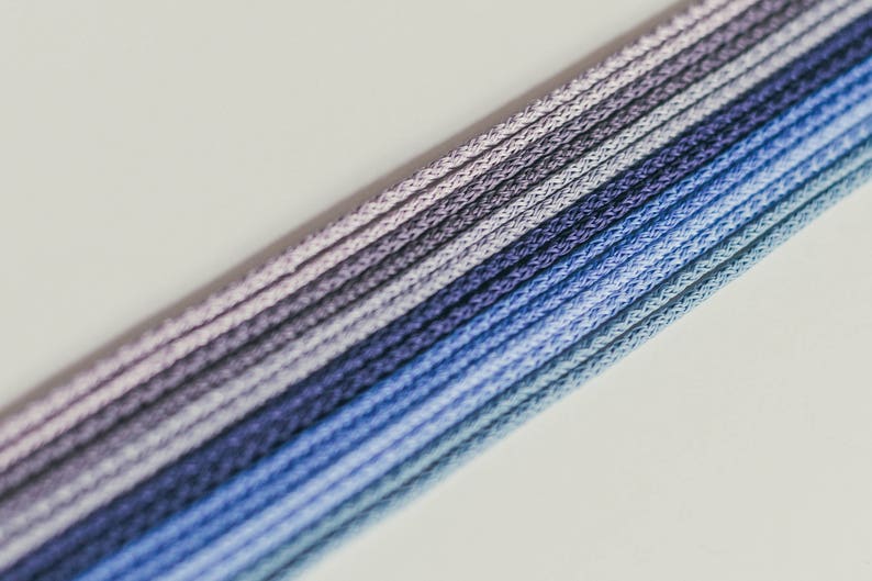 May include: Close-up of a bundle of blue, purple, and gray cords. The cords are tightly woven and arranged in parallel lines, creating a gradient effect from light to dark. The background is a plain, off-white surface.