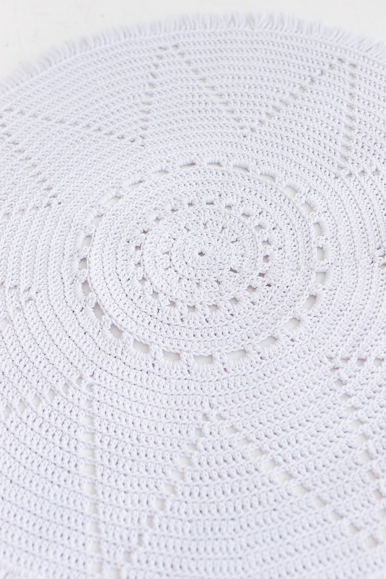 May include: A white crocheted round rug with a fringe border. The rug has a circular pattern with a centre circle and concentric circles radiating outwards.
