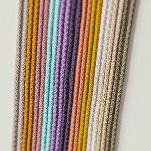 May include: Close-up of a collection of colorful, braided cords. The cords are arranged vertically, showcasing a spectrum of colors including gray, yellow, pink, teal, purple, and white. The texture of the cords is visible.