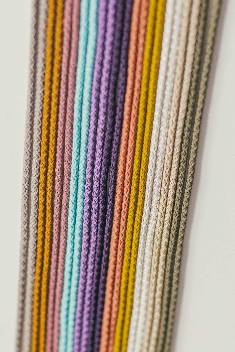 May include: Close-up of a collection of colorful, braided cords. The cords are arranged vertically, showcasing a spectrum of colors including gray, yellow, pink, teal, purple, and white. The texture of the cords is visible.