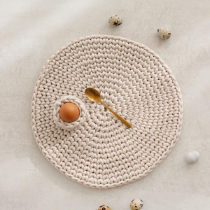 May include: A beige crocheted placemat with a small egg cup holding a brown egg and a gold spoon. Scattered around are several speckled quail eggs. The placemat is round and has a textured surface.