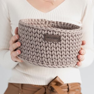 Può includere: Un cestino all'uncinetto beige fatto a mano con una forma arrotondata. Il cestino ha una piccola etichetta rettangolare in pelle con la scritta "HANDMADE". Il cestino è tenuto da una persona che indossa un maglione color crema e pantaloni marroni.