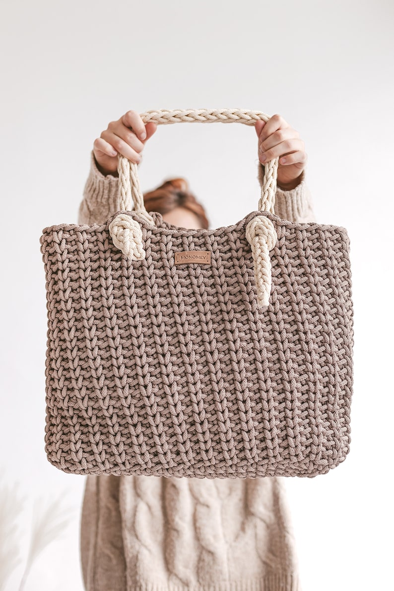 Op de afbeelding: Een bruine gehaakte tote bag met cr&egrave;mekleurige touwhandvatten en een lederen label met de tekst "MONOMEY".