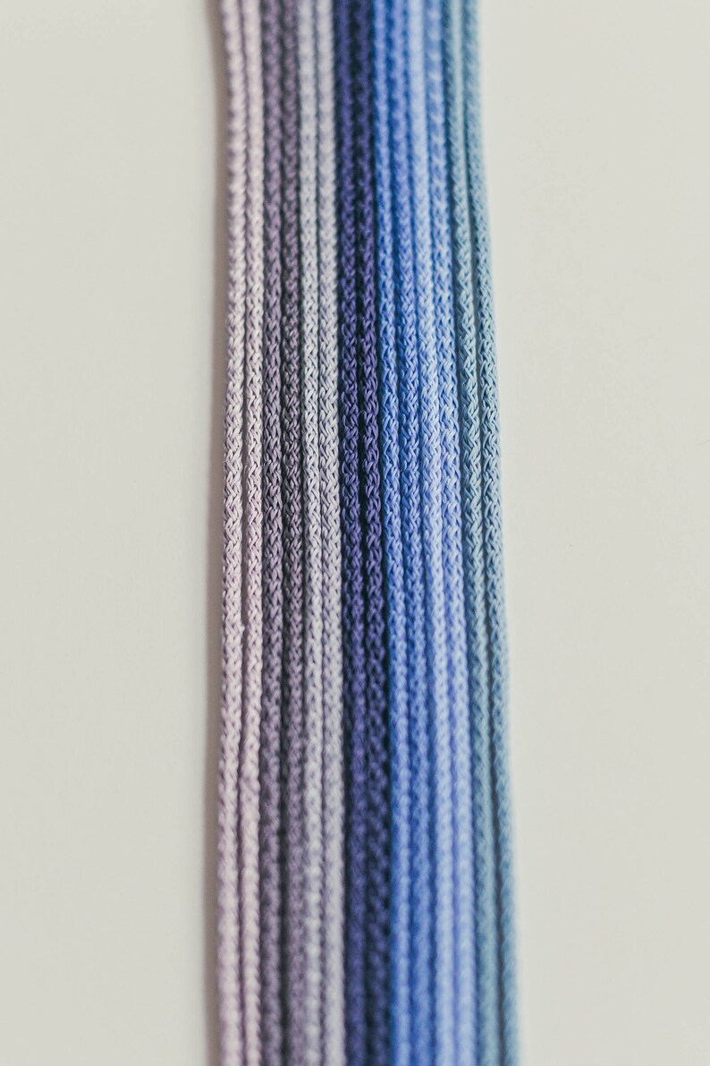 May include: Close-up of several strands of cord in a gradient of blues and purples. The cords are tightly woven, creating a textured surface. The color transitions from light lavender to deep indigo.
