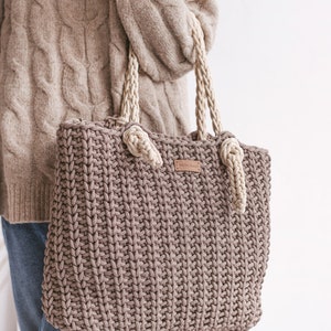 Op de afbeelding: Een bruine gehaakte tote bag met cr&egrave;mekleurige touwhandvatten. De tas heeft een lederen label met het woord "MONOCHROMY" erop.