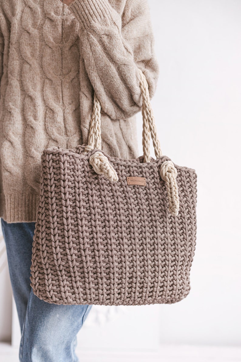 Op de afbeelding: Een bruine gehaakte tote bag met cr&egrave;mekleurige touwhandvatten. De tas heeft een lederen label met het woord "MONOCHROMY" erop.