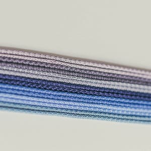 May include: Close-up of a bundle of blue, purple, and gray cords. The cords are tightly woven and arranged in parallel lines, creating a gradient effect from light blue to dark purple. The background is a plain white.