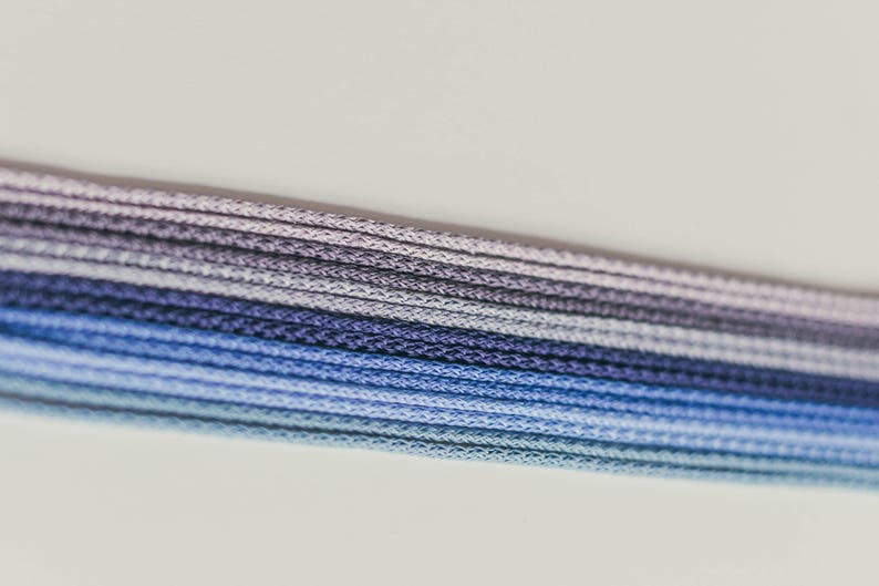 May include: Close-up of a bundle of blue, purple, and gray cords. The cords are tightly woven and arranged in parallel lines, creating a gradient effect from light blue to dark purple. The background is a plain white.