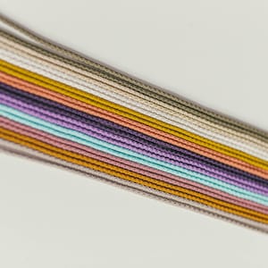 May include: A collection of colorful cords in various shades, including beige, gold, purple, and teal, arranged diagonally against a white background. The cords are tightly woven and appear to be made of a smooth material.