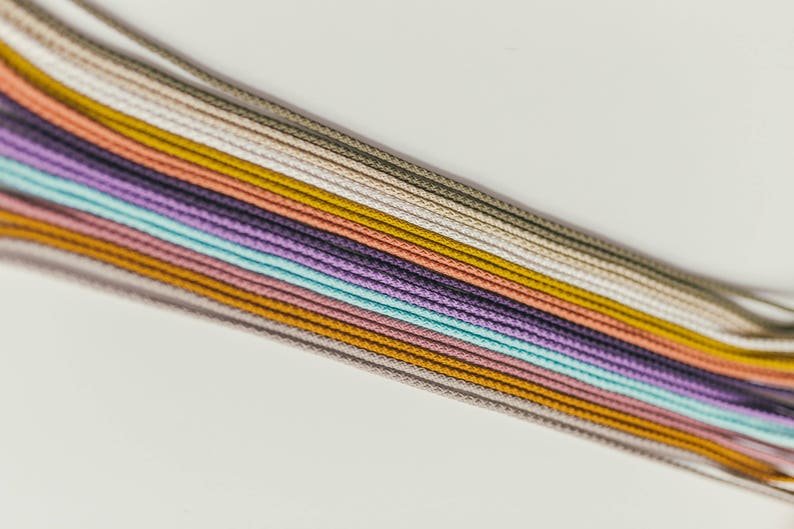May include: A collection of colorful cords in various shades, including beige, gold, purple, and teal, arranged diagonally against a white background. The cords are tightly woven and appear to be made of a smooth material.