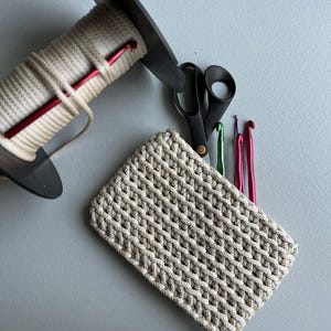 Può includere: Una collezione di strumenti e materiali per l'uncinetto, tra cui un progetto all'uncinetto beige, una bobina di filo beige, forbici nere e diversi uncinetti verdi, rosa e viola. Gli oggetti sono disposti su una superficie grigia.