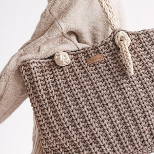 Op de afbeelding: Een bruine gehaakte tote bag met cr&egrave;mekleurige banden en een bruin leren label met de tekst "MONOMEY".